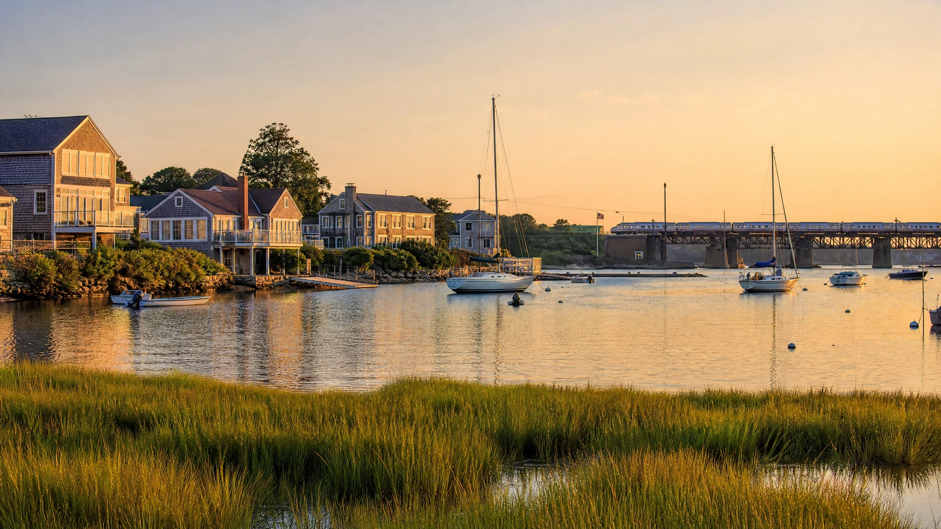 A New England coastal town harbor at golden hour with a commuter rail bridge and shingled homes along the shoreline
